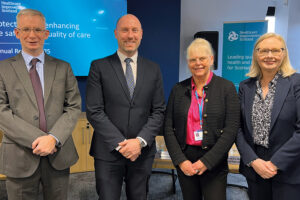 Four people standing before a screen at the Healthcare Improvement Scotland (HIS) annual review. Left to right - Robbie Pearson, Chief Executive; Neil Gray, Cabinet Secretary for Health and Social care; Evelyn McPhail, Chairman ;and Christine McLaughlin, Chief Operating Officer & Deputy Chief Executive, NHS Scotland , DG Health and Social Care
